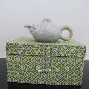 May include: A white ceramic teapot with a bird-like spout and a handle, sitting on a green and blue patterned box.