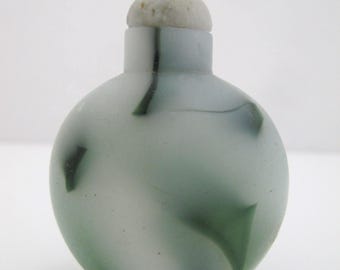 May include: A small, round, frosted glass snuff bottle featuring a translucent white base with swirling dark green and black patterns. The bottle is topped with a smooth, rounded, off-white stopper that sits securely in the narrow neck opening.