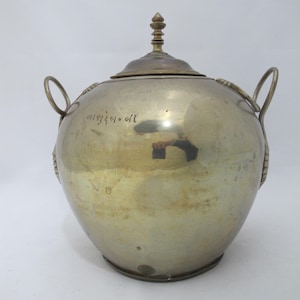 Puede incluir: Un tarro de metal plateado con tapa y dos asas. El tarro tiene un cuerpo redondo y una tapa con un adorno decorativo. Las asas son circulares. El tarro tiene algo de texto en el lateral.