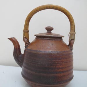 May include: A brown ceramic teapot with a bamboo handle. The teapot has a rounded body with a spout and a lid. The handle is arched and attached to the sides of the pot. The teapot is a rustic style.