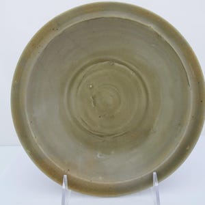 May include: A circular ceramic plate with a muted olive-green glaze. The surface features concentric circular indentations radiating from the center, with subtle variations in color and texture across the glazed finish and a wide, flat rim.