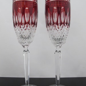 May include: Two tall crystal champagne flutes featuring a deep red upper bowl with vertical cut patterns and a clear diamond-cut base. Each glass has a slender faceted stem and a circular foot with a radial starburst design etched into the glass.