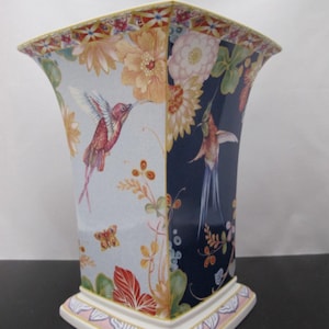 May include: A decorative ceramic vase with a square base and flared top. The vase features a colourful floral and hummingbird design. The background is divided into light blue and navy blue sections. The base and rim have decorative patterns.