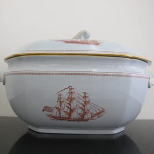 May include: A white porcelain soup tureen with a lid, featuring a gold rim and handles. The tureen is decorated with a red and white ship illustration. The lid also has a ship illustration and a gold trim.