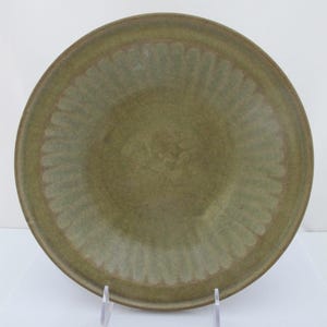 May include: A round, olive-green ceramic bowl featuring a wide, flat rim with a repeating, embossed vertical fluted pattern. The interior of the bowl has a smooth, matte finish with subtle color variations, and the edges are slightly rounded.