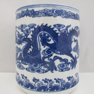 May include: White ceramic cylindrical vase with a blue dragon design. The vase features a detailed illustration of a dragon amidst clouds, with wave patterns at the base and a cloud border at the top. The design is in shades of blue.