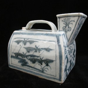 May include: A rectangular ceramic vessel with a blue and white hand-painted finish. The object features a top handle and a wide, angled spout. The side panels are decorated with blue botanical illustrations, including stylized plants and foliage.