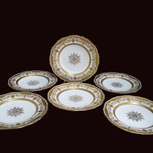 May include: A set of six white porcelain plates with gold rims and intricate gold floral and geometric patterns. The plates have a scalloped edge design. The center of each plate features a detailed gold starburst design.