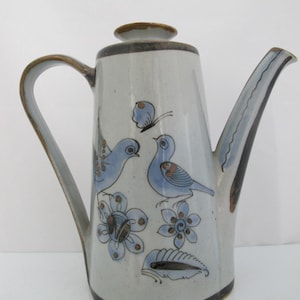 May include: A ceramic teapot with a light gray speckled glaze, featuring hand-painted blue birds, a butterfly, and floral motifs in blue and brown. The teapot has a tall, tapered body, a curved handle, a long spout, and a matching lid with brown trim.