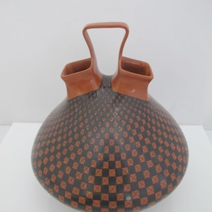 May include: A terracotta-colored ceramic vessel with a rounded body and two distinct openings connected by a central arched handle. The lower portion of the vessel features a repeating black and terracotta checkered pattern with small cross marks.