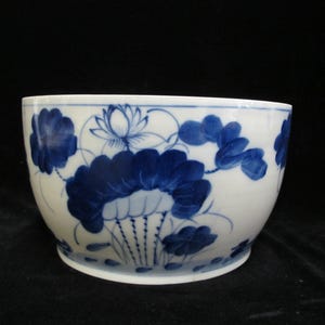 Op de afbeelding: Een witte keramische kom met een blauw bloemmotief. De kom heeft een herhalend patroon van lotusbloemen en bladeren. Het ontwerp is met de hand geschilderd in een traditionele stijl, met een donkerblauwe kleur tegen de witte achtergrond.