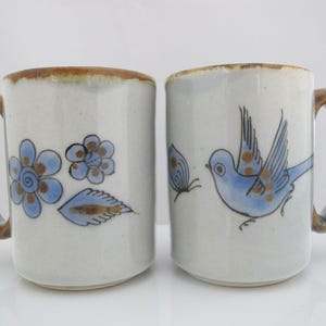 May include: Two ceramic mugs with a speckled off-white glaze and brown rim accents. One mug features blue hand-painted flowers and a leaf, while the other displays a blue bird and a butterfly. Both mugs have curved handles with brown detailing.