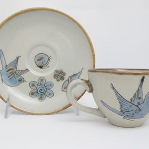 May include: A ceramic cup and saucer set featuring a hand-painted light blue bird with brown accents on the cup and a matching saucer decorated with two blue birds, a blue flower, and a small brown leaf motif, all set against a cream background.