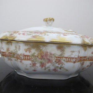 May include: A white porcelain serving dish with a lid, featuring a floral pattern in shades of pink, green, and gold. The dish has a scalloped edge and gold trim.