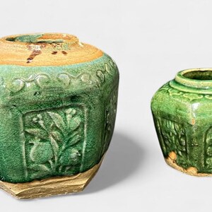 May include: Two antique green ceramic jars with floral relief patterns. The larger jar has a wide, octagonal shape with a tan rim. The smaller jar is a similar design, but smaller. Both jars have a weathered appearance.