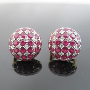 May include: A pair of round stud earrings. Each earring has a spherical design with a grid pattern of vibrant red gemstones, interspersed with small, sparkling white diamonds. The earrings are set in a silver metal.