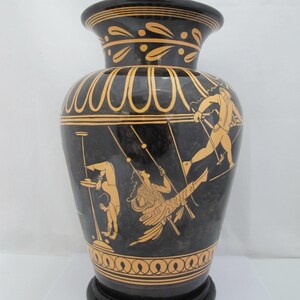 May include: A black ceramic vase featuring orange-toned illustrations of figures in motion. The neck and base are decorated with repeating leaf and scroll patterns. The central body depicts individuals performing acrobatic feats with poles.