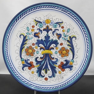 May include: A round, decorative ceramic plate with a white background and a blue border. The plate features a central design of blue, orange, and yellow floral and leaf motifs. The border has a blue and white striped pattern.