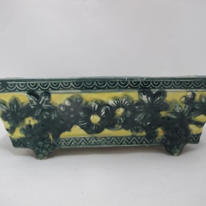以下が含まれることがあります： 黄色い内装と濃い緑色の花とブドウのつるのデザインが施された長方形のセラミックプランター。 プランターはスカラップエッジで、3つの小さな足で支えられています。 家のための装飾的な作品。