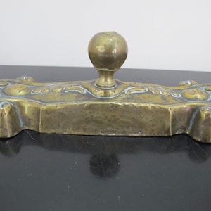 May include: A brass inkwell with an ornate design and a round knob on top.