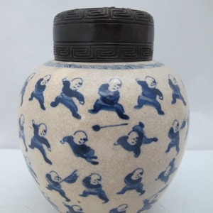 Puede incluir: Un tarro de jengibre de cerámica con tapa de madera oscura. El tarro es de color crema con un esmalte craquelado y decorado con figuras azules. La tapa tiene un patrón geométrico.