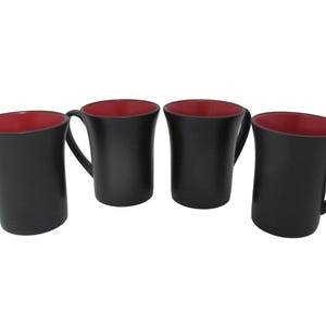 Könnte beinhalten: Vier schwarze Keramikbecher mit rotem Innenraum. Jeder Becher hat einen gebogenen Henkel und eine leicht ausgestellte Form. Die Becher sind in einer Reihe vor weißem Hintergrund angeordnet.