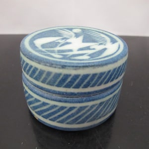 Peut inclure: Petite boîte à bijoux ronde en céramique bleue et blanche. Le couvercle présente un motif blanc d'oiseau et d'autres formes. Les côtés de la boîte ont un motif à rayures diagonales. La boîte est posée sur une surface sombre.