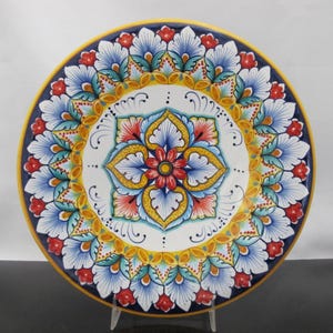 Op de afbeelding: Een decoratieve keramische plaat met een levendig bloemenontwerp. De plaat heeft een wit midden met een centraal bloemmotief in geel, rood en blauw. De rand is versierd met blauwe en rode bloempatronen, geplaatst tegen een gele en marineblauwe rand.