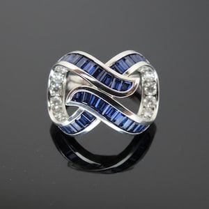 May include: A silver ring with an intertwined design. The ring features blue rectangular gemstones and clear round gemstones. The ring is displayed on a reflective black surface.