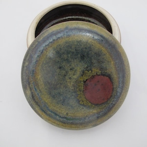 May include: A ceramic jar with a lid. The lid is round with a dark red circle in the center, surrounded by green, blue, and yellow hues. The jar's rim is off-white, and the interior is dark brown.