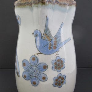 Op de afbeelding: Een keramieken kan met een wit geglazuurde afwerking, voorzien van handgeschilderde lichtblauwe vogels en blauwe bloemen met bruine harten. De rand heeft een gevlekt bruin en blauw glazuur en de kan heeft een licht taps toelopende vorm.