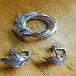 May include: A silver tone brooch and matching earrings featuring a leaf design. The brooch is a circle with three rows of metal and a leaf design on the side. The earrings are clip-on and feature a single leaf design.