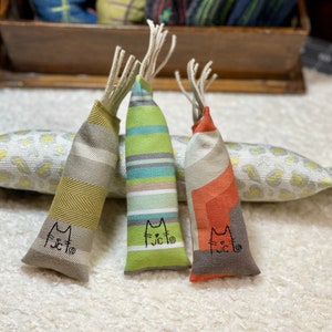 Puede incluir: Tres juguetes para gatos con forma de pez. Los juguetes están hechos de tela y tienen diferentes patrones. Todos los juguetes están etiquetados con la palabra "fucto" en bordado negro.