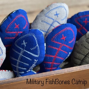 Puede incluir: Un conjunto de seis juguetes para gatos con forma de espinas de pescado. Los juguetes están hechos de tela y tienen un esquema de colores rojo, blanco y azul. Los juguetes están en una caja de madera con el texto "Military FishBones Catnip Toy" impreso en la parte delantera.