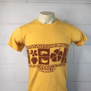 May include: A yellow short-sleeved t-shirt with a brown print of Hawaiian-themed imagery, including a hibiscus flower, footprints, and a seashell. The print also includes the word "HAWAII" in brown text.