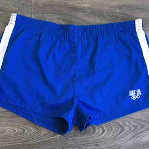 Può includere: Pantaloncini sportivi blu reale con strisce laterali bianche. I pantaloncini presentano un elastico in vita e un piccolo logo con i cerchi olimpici. Uno stile vintage, perfetto per la corsa o per l'abbigliamento casual.