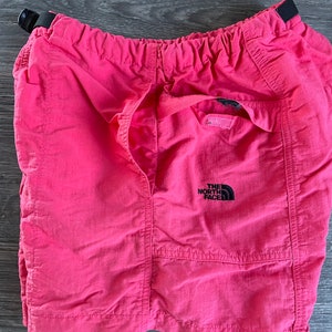 May include: Pink nylon shorts with an elastic waistband and a black The North Face logo on the right leg.