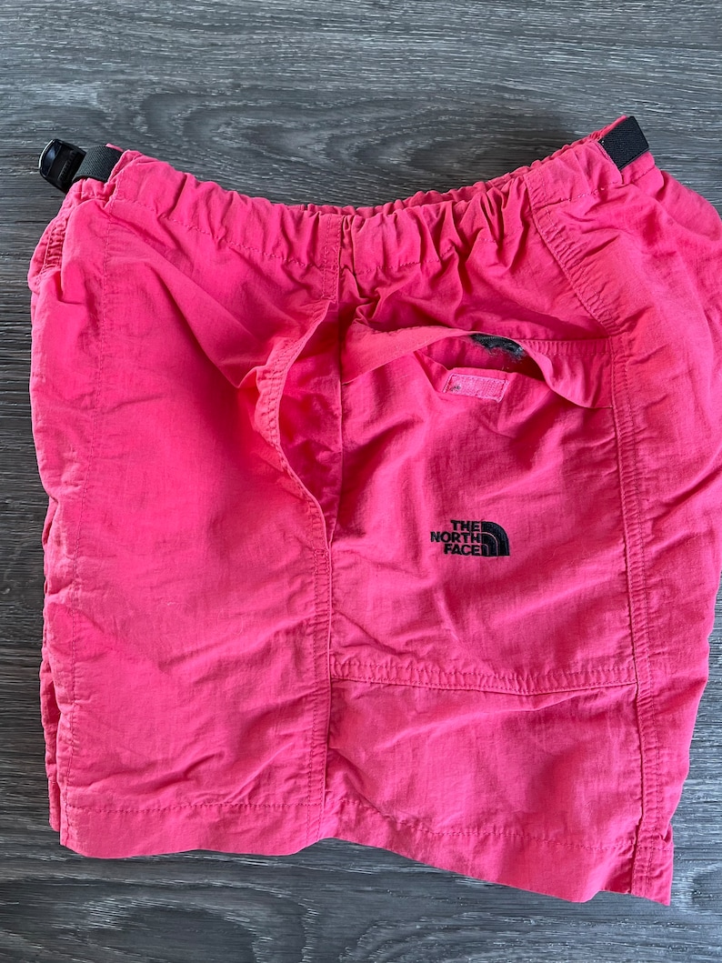 May include: Pink nylon shorts with an elastic waistband and a black The North Face logo on the right leg.