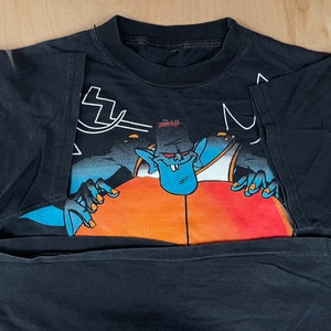 May include: Black t-shirt featuring a cartoon character in a blue and orange outfit holding a basketball. The character has blue skin and a large mouth with sharp teeth.