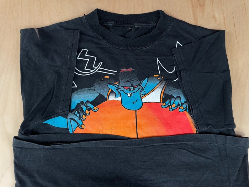 May include: Black t-shirt featuring a cartoon character in a blue and orange outfit holding a basketball. The character has blue skin and a large mouth with sharp teeth.
