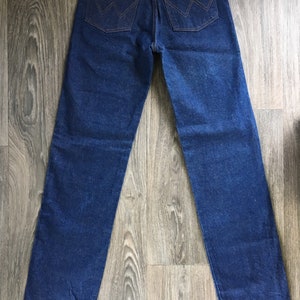 Puede incluir: Un par de pantalones vaqueros de mezclilla azul oscuro con un parche de cuero en el bolsillo trasero que dice "Wrangler".