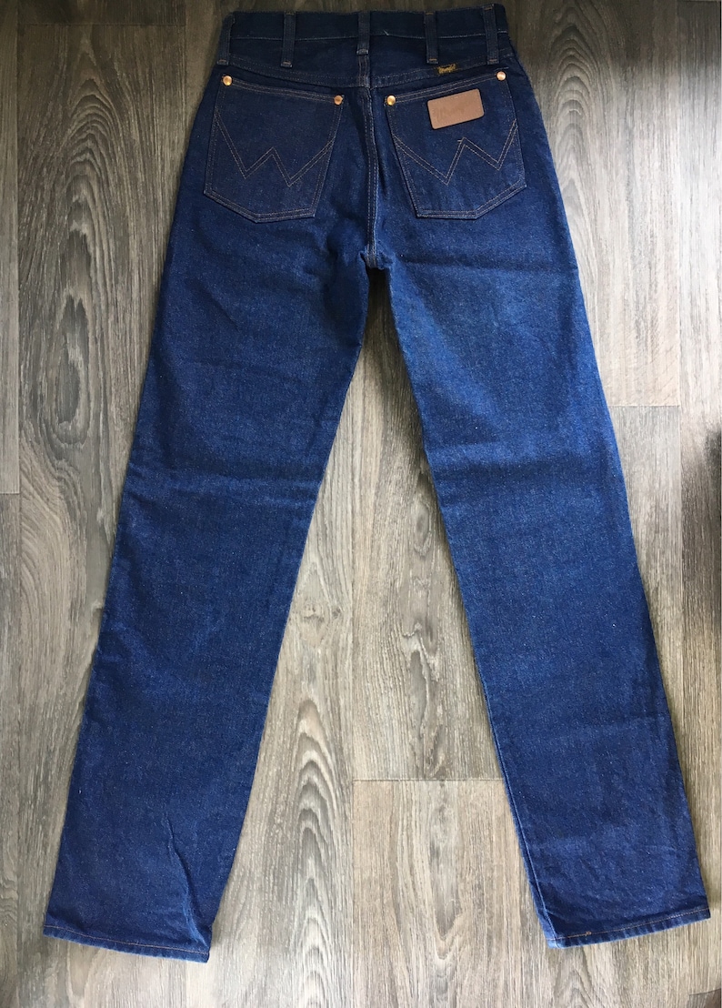 Puede incluir: Un par de pantalones vaqueros de mezclilla azul oscuro con un parche de cuero en el bolsillo trasero que dice "Wrangler".