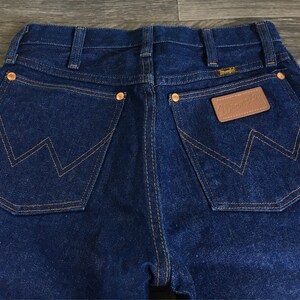 Puede incluir: Un par de pantalones vaqueros de mezclilla azul oscuro con un parche de cuero marr&oacute;n que dice "Wrangler" en el bolsillo trasero derecho. Los pantalones tienen un estilo cl&aacute;sico de cinco bolsillos con pierna recta.