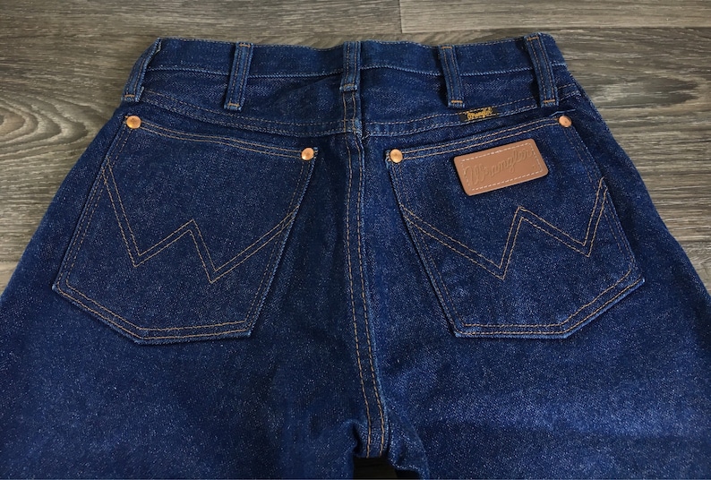 Puede incluir: Un par de pantalones vaqueros de mezclilla azul oscuro con un parche de cuero marr&oacute;n que dice "Wrangler" en el bolsillo trasero derecho. Los pantalones tienen un estilo cl&aacute;sico de cinco bolsillos con pierna recta.