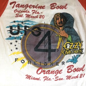 May include: A white t-shirt with a red and white baseball sleeve design. The shirt features a graphic of a circle with the number 4 inside, the words "UFO 4 FOREIGNER" and a cartoon image of a man with a sword. The text "Tangerine Bowl Orlando, Fla. - Sat. March 20" and "Orange Bowl Miami, Fla. - Sun. March 21" are printed on the shirt.