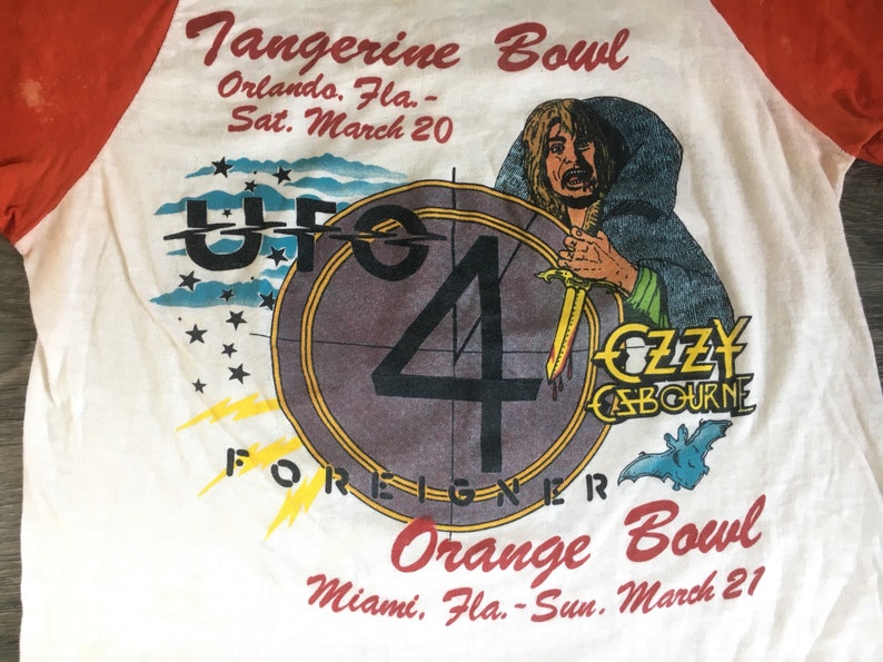 May include: A white t-shirt with a red and white baseball sleeve design. The shirt features a graphic of a circle with the number 4 inside, the words "UFO 4 FOREIGNER" and a cartoon image of a man with a sword. The text "Tangerine Bowl Orlando, Fla. - Sat. March 20" and "Orange Bowl Miami, Fla. - Sun. March 21" are printed on the shirt.