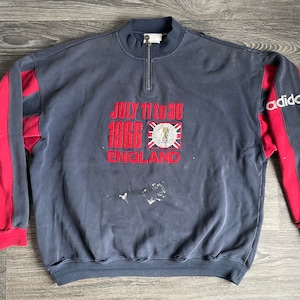 May include: A navy blue sweatshirt with red sleeves and a white Adidas logo on the right sleeve. The front of the sweatshirt has an embroidered design that says "JULY 11 to 30 1966 ENGLAND" with a crest and a Union Jack flag.