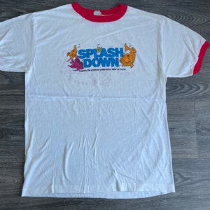 May include: White t-shirt with a red ringer. The shirt has a graphic of a cartoon fish with the text "Splash Down" and "Explore the greatest underwater show on earth".