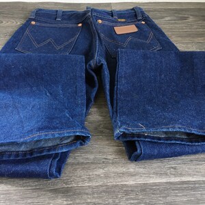 Puede incluir: Un par de pantalones vaqueros de mezclilla azul oscuro con un parche de cuero marr&oacute;n en el bolsillo trasero. Los pantalones vaqueros est&aacute;n colocados sobre una superficie de madera.