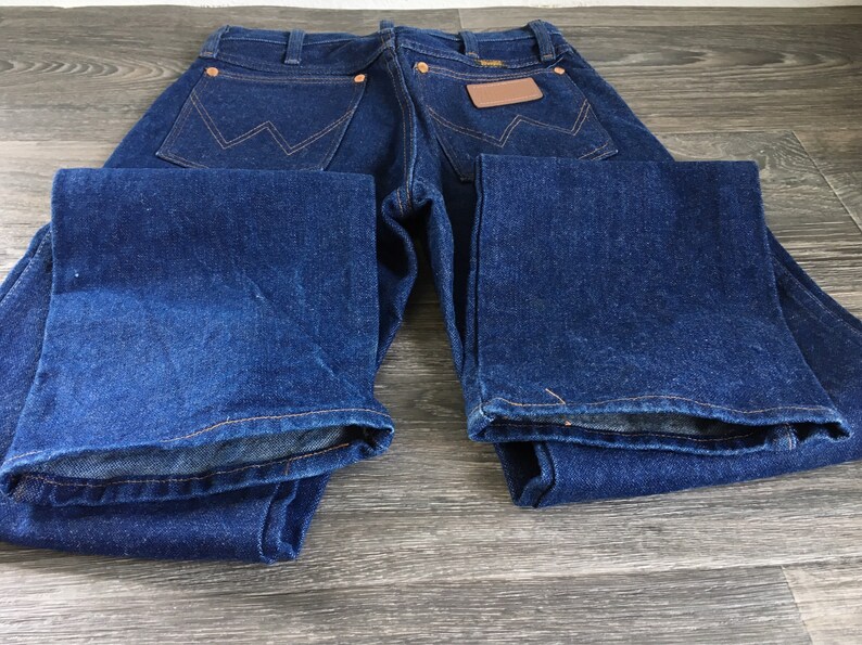 Puede incluir: Un par de pantalones vaqueros de mezclilla azul oscuro con un parche de cuero marr&oacute;n en el bolsillo trasero. Los pantalones vaqueros est&aacute;n colocados sobre una superficie de madera.